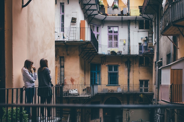 Two young women stand in patio together and look at it. Colorful windows in old city or town. Travel together