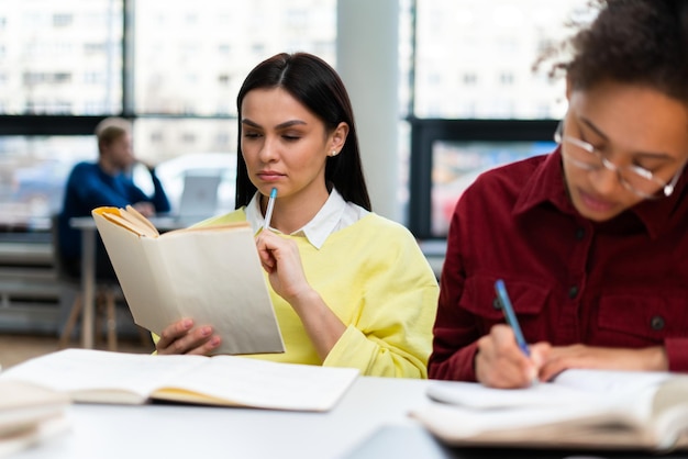 Two young women working together with concentration while sitting at table Young university student reading book while her classmate writing something