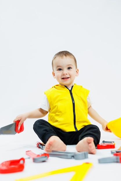 A twoyearold boy in a yellow vest sits and plays with plastic construction tools on a white background Toys for small children