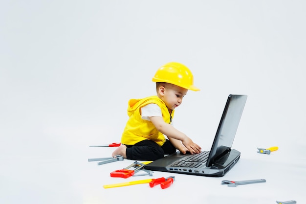 A twoyearold boy in a yellow vest on a white background sits with a laptop and plays with plastic construction tools Toys for small children