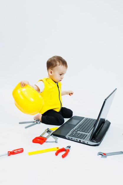 A twoyearold boy in a yellow vest on a white background sits with a laptop and plays with plastic construction tools Toys for small children