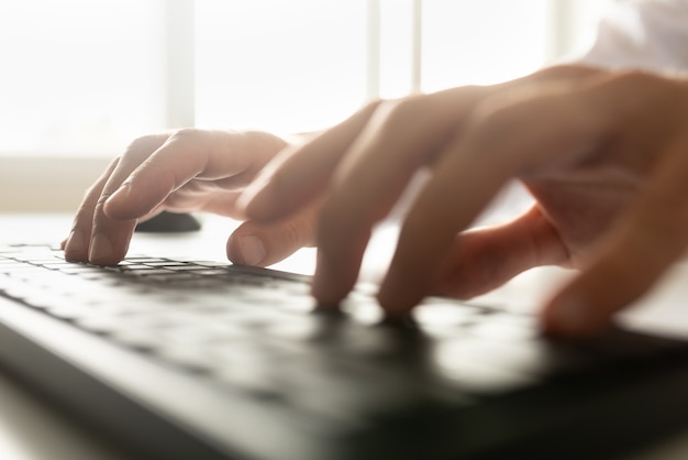 Typing on a computer keyboard with a bright light flare coming in through a window over his hands.