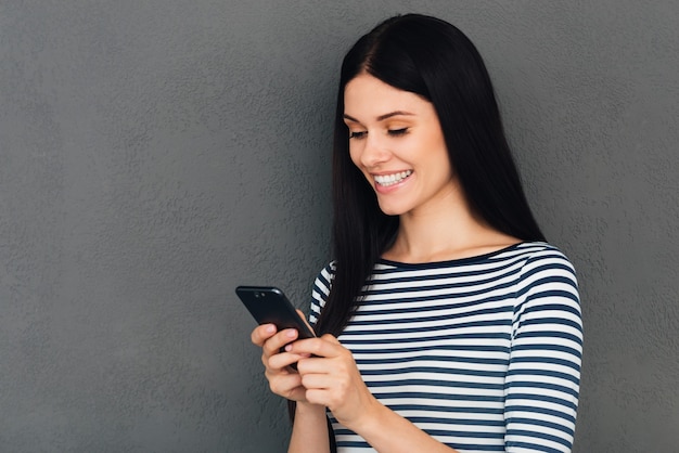 Typing message to friend. Attractive young woman holding smart phone and looking at it with smile standing