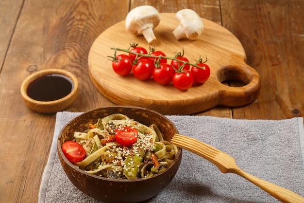 Udon with vegetables and mushrooms on the table in a wooden plate next to champignons and tomatoes on a plank