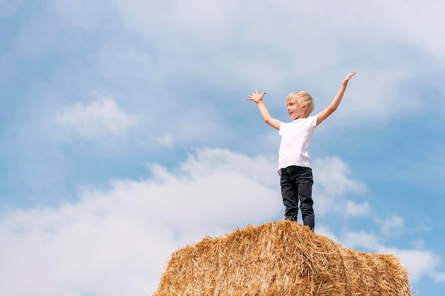 Ukrainian fairhaired stands on the haystack with his hands up against the blue sky and clouds Weekend in the village