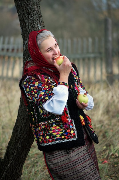 Photo ukrainian girl in national hutsul clothes leans against the trunk of an apple tree and eats an apple