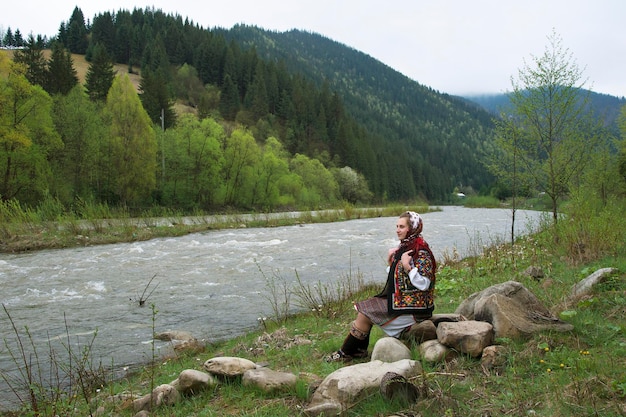 Ukrainian hutsul woman in authentic national costume sitting on a stone on the bank of the river.