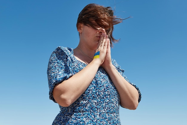 Ukrainian woman with a flag symbol on her hand prays against the blue sky stop war