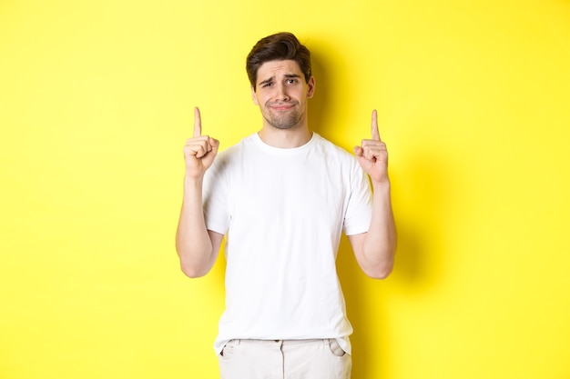 Unamused handsome guy frowning, pointing fingers up at something bad, standing skeptical against yellow background