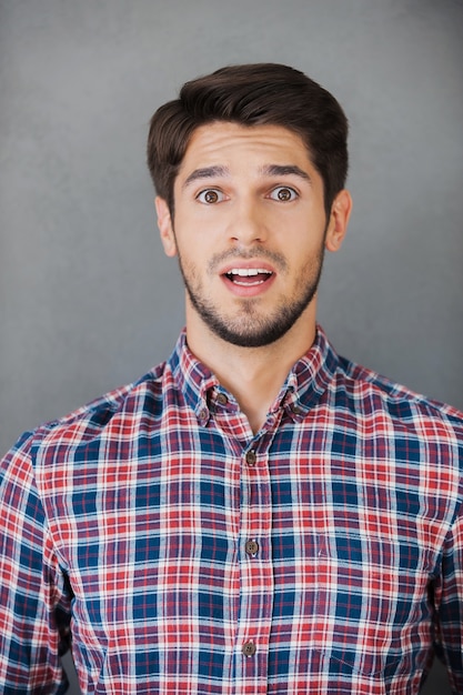 Unbelievable! Surprised young man in shirt staring at camera while standing against grey background