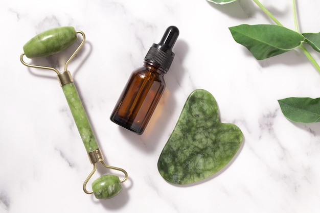 Unbranded serum bottle face roller and guasha with leaves on marble background Jade face roller for facial massage therapy Anti age lifting and toning treatment