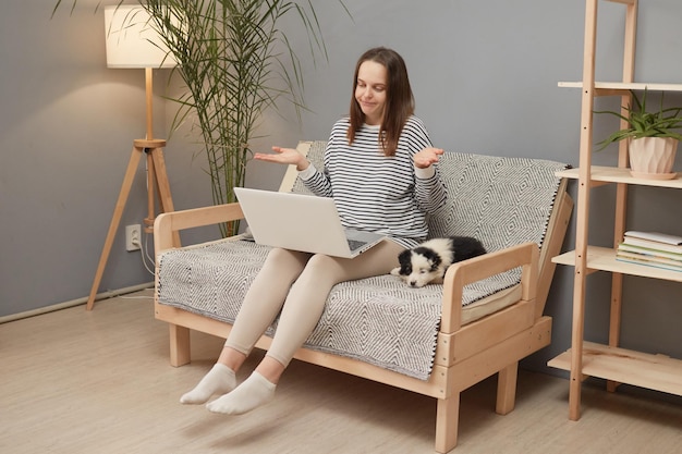 Uncertain Caucasian woman wearing striped shirt sitting on sofa with her puppy dog having video call