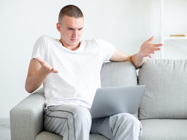 Uncertain situation Hesitating man Online meeting Confused casual guy sitting sofa looking laptop with shrugging hands in light room interior