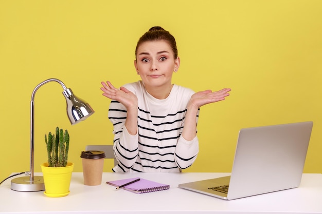 Uncertain woman office worker in striped shirt shrugging shoulders sitting on laptop at workplace