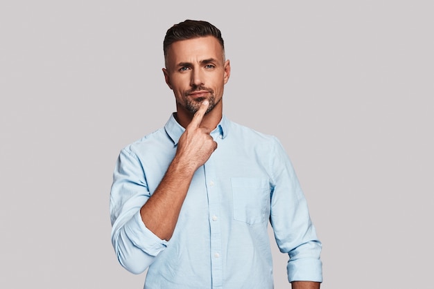 Uncertainty. Thoughtful young man keeping hand on chin and looking at camera while standing against grey background