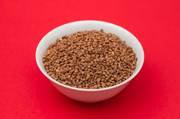 Uncooked buckwheat in small white bowl on red