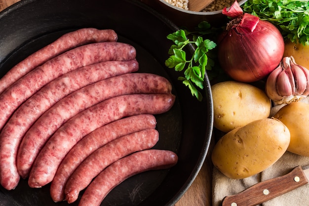 Uncooked raw pork sausages in iron cast frying pan vegetables herbs on kitchen table dinner