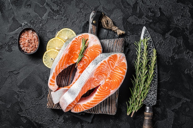 Uncooked Raw Salmon Fish Steaks on butcher board with herbs. Black background. Top view.