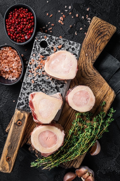 Uncooked Raw veal Marrow bones on butcher board with meat cleaver. Black background. Top view.