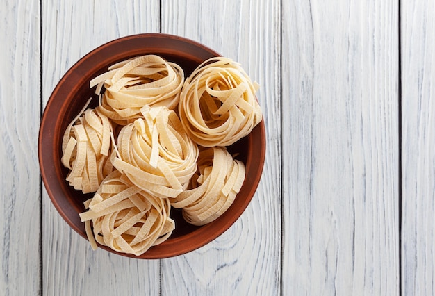 Uncooked tagliatelle pasta in ceramic bowl on white wooden background