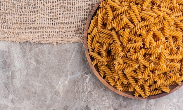 Uncooked whole wheat fusilli on a wooden plate on a burlap on the marble surface