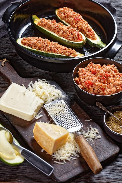 Uncooked zucchini boats with ground meat, chopped tomatoes, and cheese in a black baking dish on a wooden table with ingredients, italian cuisine