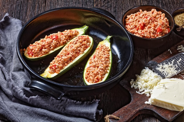 Uncooked zucchini boats with ground meat, chopped tomatoes, and cheese in a black baking dish on a wooden table with ingredients, italian cuisine