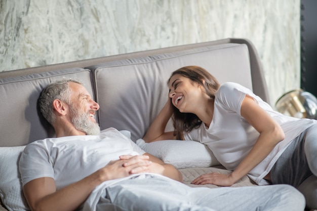 Understanding. Joyful cheerful gray-haired man and dark-haired woman looking at each other in pajamas on bed