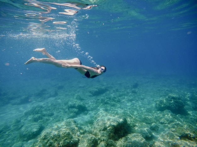 Underwater side view of attractive young cheerful girl diving with goggles and looking at the camera.