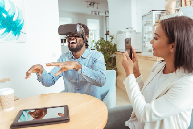 Unforgettable emotions. Beautiful upbeat woman sitting in the coffeehouse next to her best friend and filming him while he testing his new VR headset