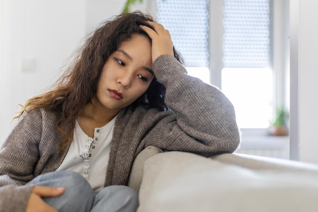 Unhappy Asian woman on sofa crying Lonely sad woman deep in thoughts sitting daydreaming or waiting for someone in the living room with a serious expression sitting on couch