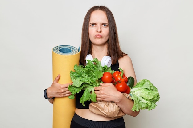 Unhappy Caucasian woman holding organic vegetables standing isolated over white background looking at camera with pout lips offended expression has workout on mat