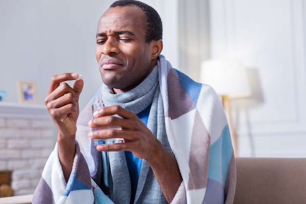 Unhappy cheerless ill man holding a pill and looking at it while not wanting to take it