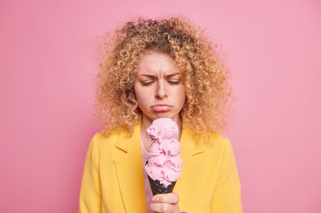 Unhappy curly haired European woman looks sadly at gelato feels temptation cannot afford herself eating high calorie food dressed in yellow clothing isolated over pink wall. Tasty ice cream