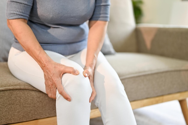 Unhappy elderly Asian woman sits on sofa in her living room suffering from knee pain