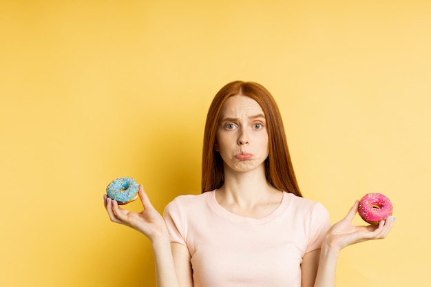 Photo unhappy frustrated slim ginger young woman on diet holding two color donuts, can't eat delicious confectionery, isolated over yellow background. junk food, dieting, weight loss concept.