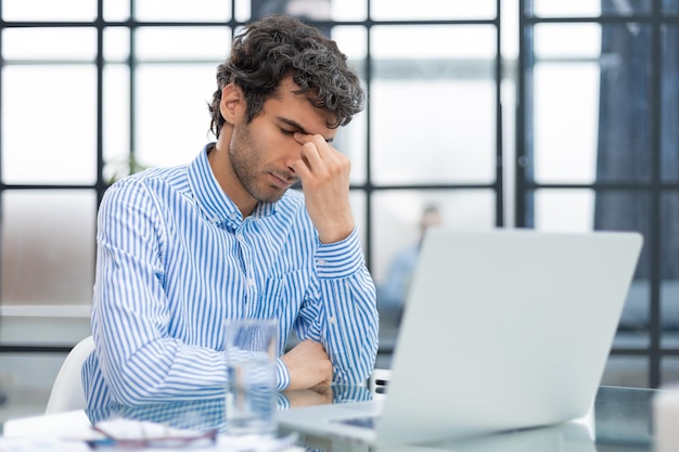 Unhappy frustrated young male holding head by hands sitting with computer behind desk at office