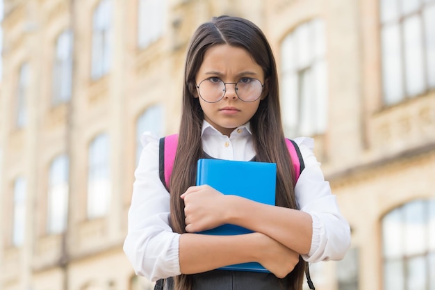 Unhappy little kid in eyeglasses hold study book in schoolyard outdoors back to school