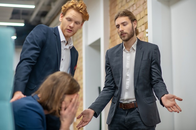 Unhappy man sitting covering his face with hands and two supportive colleagues standing nearby indoors