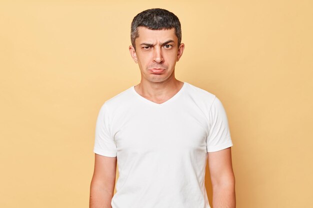 Unhappy offended man wearing white casual tshirt standing isolated over beige background looking at camera with pout lips expressing sorrow and sadness