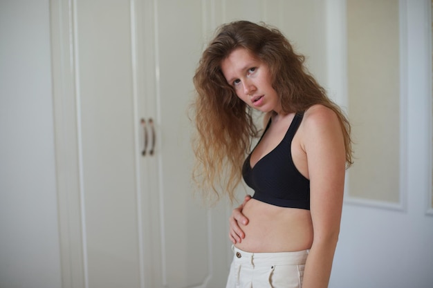 Unhappy sad upset young woman with fat on her stomach girl worried about body fat touching her belly standing at home near closet