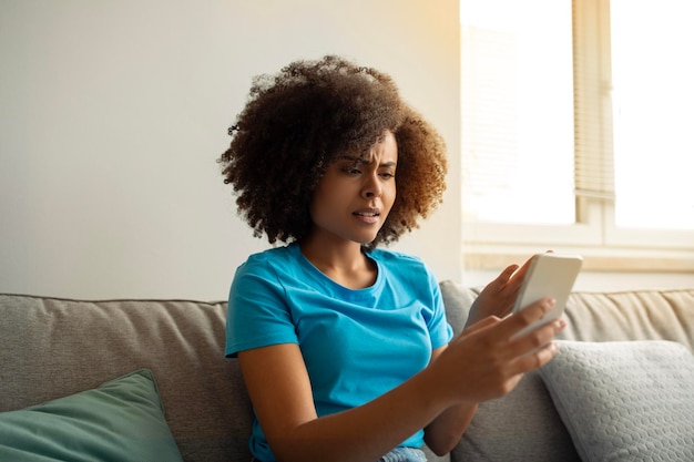 Unhappy sad young black curly woman reads message on smartphone find mistake in living room