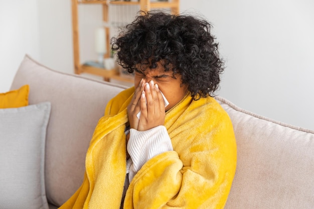 Unhappy sick young african american woman with cold sitting on sofa at home girl covering with blank