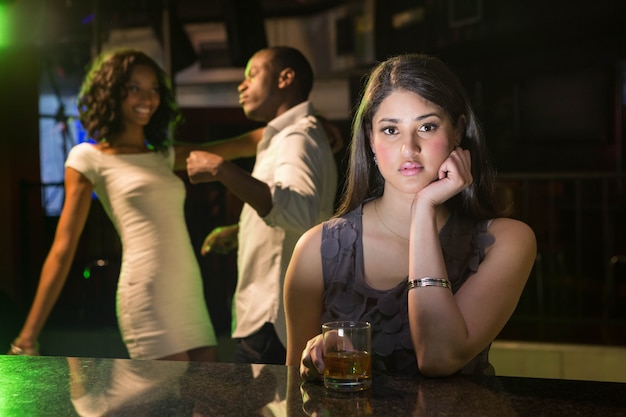 Unhappy woman sitting at bar counter and couple dancing behind her in bar