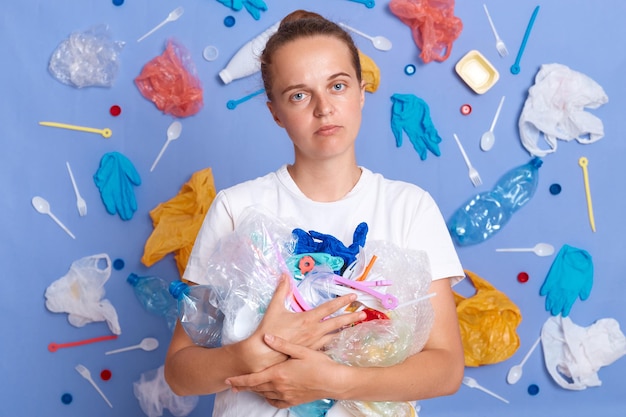 Unhappy woman wearing white shirt isolated on blue wall with litter around holding garbage looking at camera wants to save our planet reduce waste pollution