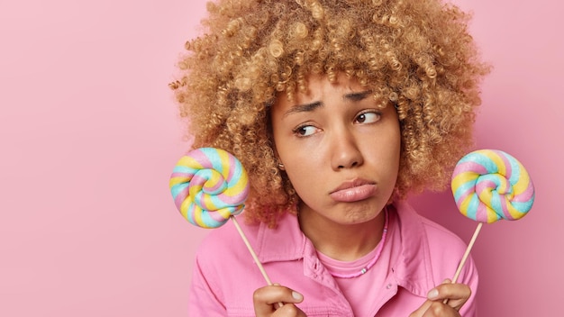 Unhappy woman with curly hair holds two colorful lollipops has sad expression keeps to diet purses lips wears fashionable jacket isolated over pink background empty space for your promotion