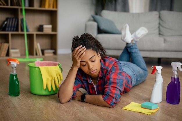 Unhappy young black female in rubber gloves lies on floor with cleaning supplies in living room interior