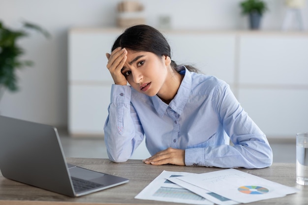 Unhappy young hindu female manager presses hand to head and looks at laptop suffers from headache in office