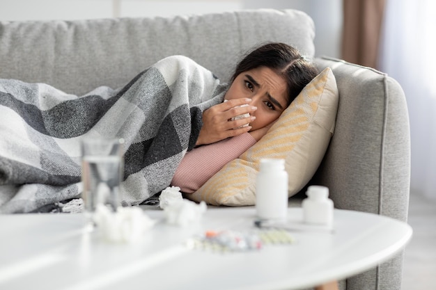 Unhappy young indian female sneezes in napkin blows nose suffers from fever and flu lies on sofa