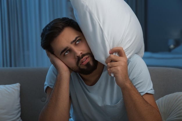 Unhappy young man covering ears in living room at night Noisy neighbours